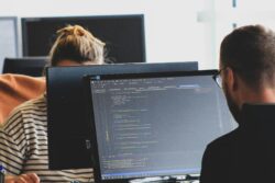 Man in black shirt sitting beside a flat screen computer monitor with code on it.