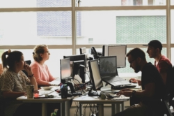 Four colleagues collaborating at a shared workspace, focused on their monitors. The group includes individuals in a red top, a striped top, a black t-shirt and glasses, and a baseball cap and red t-shirt.