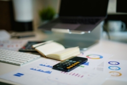 An open iPhone calculator surrounded by papers, charts, and a laptop on a stand, suggesting focused data analysis or financial planning.