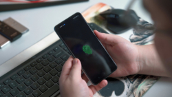 A person holding a smartphone displaying a green fingerprint icon on the screen, representing biometric authentication as part of digital payment solutions. A keyboard and mouse are visible in the background on a desk.