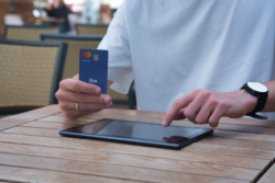 Close-up of a person in a white t-shirt holding a Fire business debit card in one hand while tapping on a tablet placed on a table with the other hand, illustrating the concept of making an online purchase.