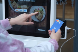 Close-up of a person in a pink jumper using a Fire business debit card to make a purchase at a touchscreen. They select a food item on the screen and then tap the card on a POS terminal to complete the payment.