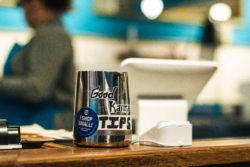 A stainless steel tip jar with "Good Karma” and “TIPS" written on it sits on a wooden counter near a point-of-sale system. The background is blurred, showing a person in an apron working in a coffee shop.