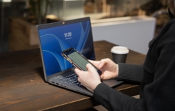 A person in a dark jumper sits at a wooden table using a smartphone, with a laptop open in front of them. A takeaway coffee cup is placed beside the laptop.