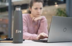 A grey Fire-branded water bottle sits on a desk in sharp focus, with a blurred woman in a pink jumper working on a laptop in the background. A notebook and pen are also visible on the desk.