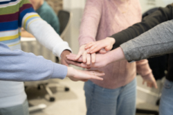 Close-up of the hands of a group of people standing in a circle, placing their hands together in a stack to symbolise teamwork and unity. Their casual clothing suggests a relaxed and collaborative culture.