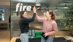 Two women smiling and giving each other a high-five in front of the glass wall of Fire’s office, which displays the Fire logo and the tagline “Trusted. Integrated. Payments.” Both are casually dressed in a modern office setting, suggesting a friendly and collaborative workplace culture.