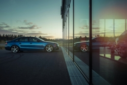 A blue sedan parked outside a modern glass building at sunset, with the car and surrounding landscape reflected clearly in the large windows. The scene suggests a car dealership setting, highlighting the operational environment of multi-location businesses.