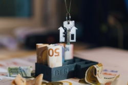 A house-shaped keychain with a key hangs above a model building filled with rolled euro banknotes. Additional notes and coins are scattered on printed financial documents in the background, symbolising property payments, financial oversight, and real estate management.