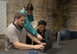 Three colleagues collaborating at a table, smiling and discussing work on a laptop in a modern office with stone walls.
