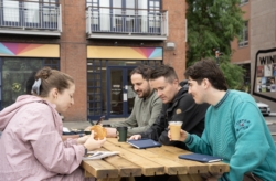 Four people sit around a wooden picnic-style table outside a coffee shop. One person is holding a pastry, another has a coffee cup, and two have notebooks on the table.