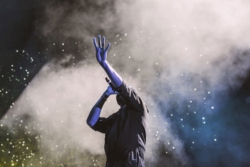 Performer holding a microphone on stage with one arm raised against a backdrop of smoke and lights.