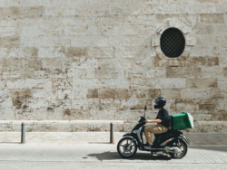 A delivery worker rides a scooter with a green insulated box on the back, passing a stone wall with a small round window.