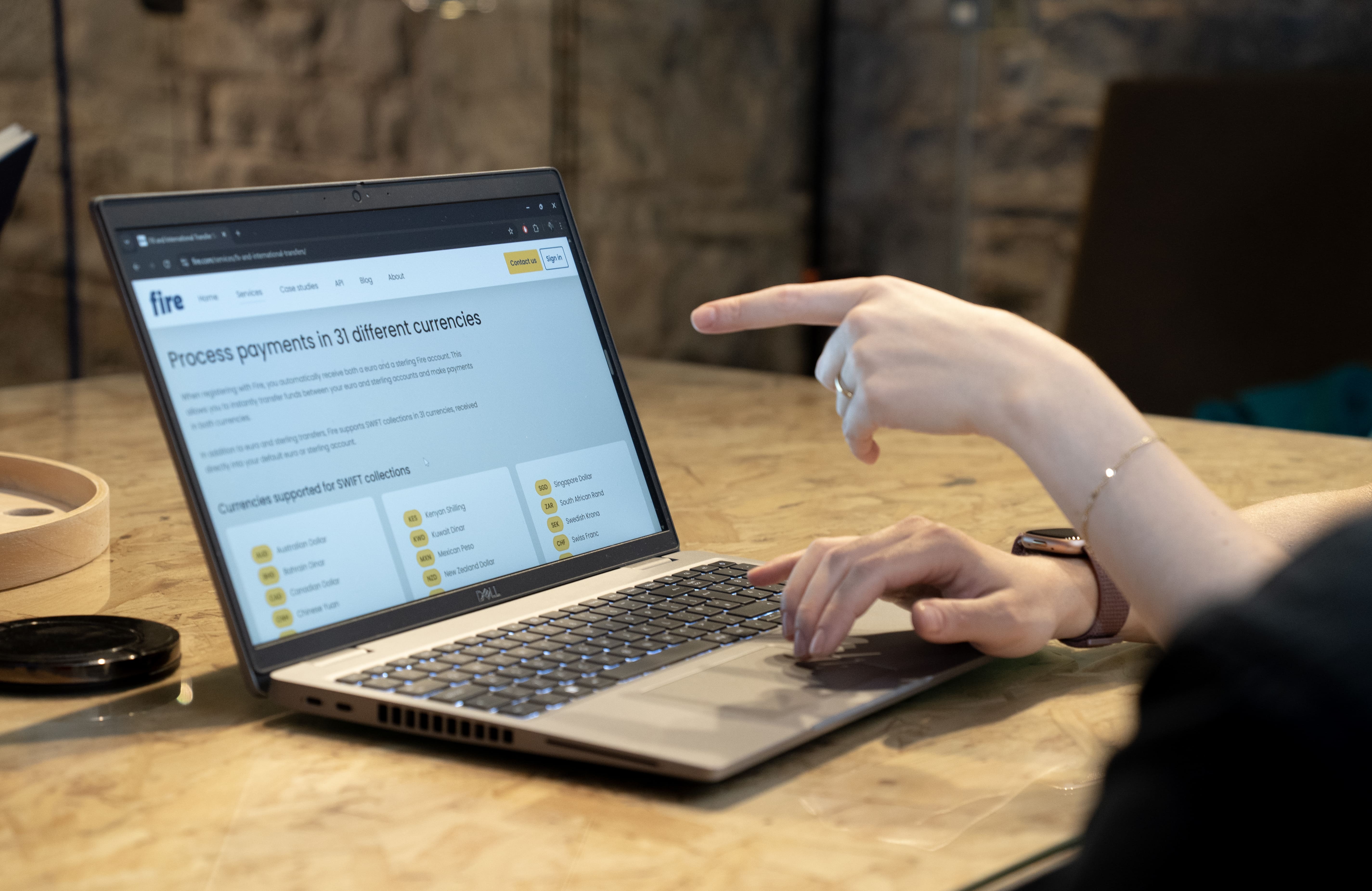 Close-up of a laptop on a wooden desk displaying Fire’s website page on FX services, with the title “Process payments in 31 different currencies” visible, with one hand pointing at the screen and the other resting on the keyboard.