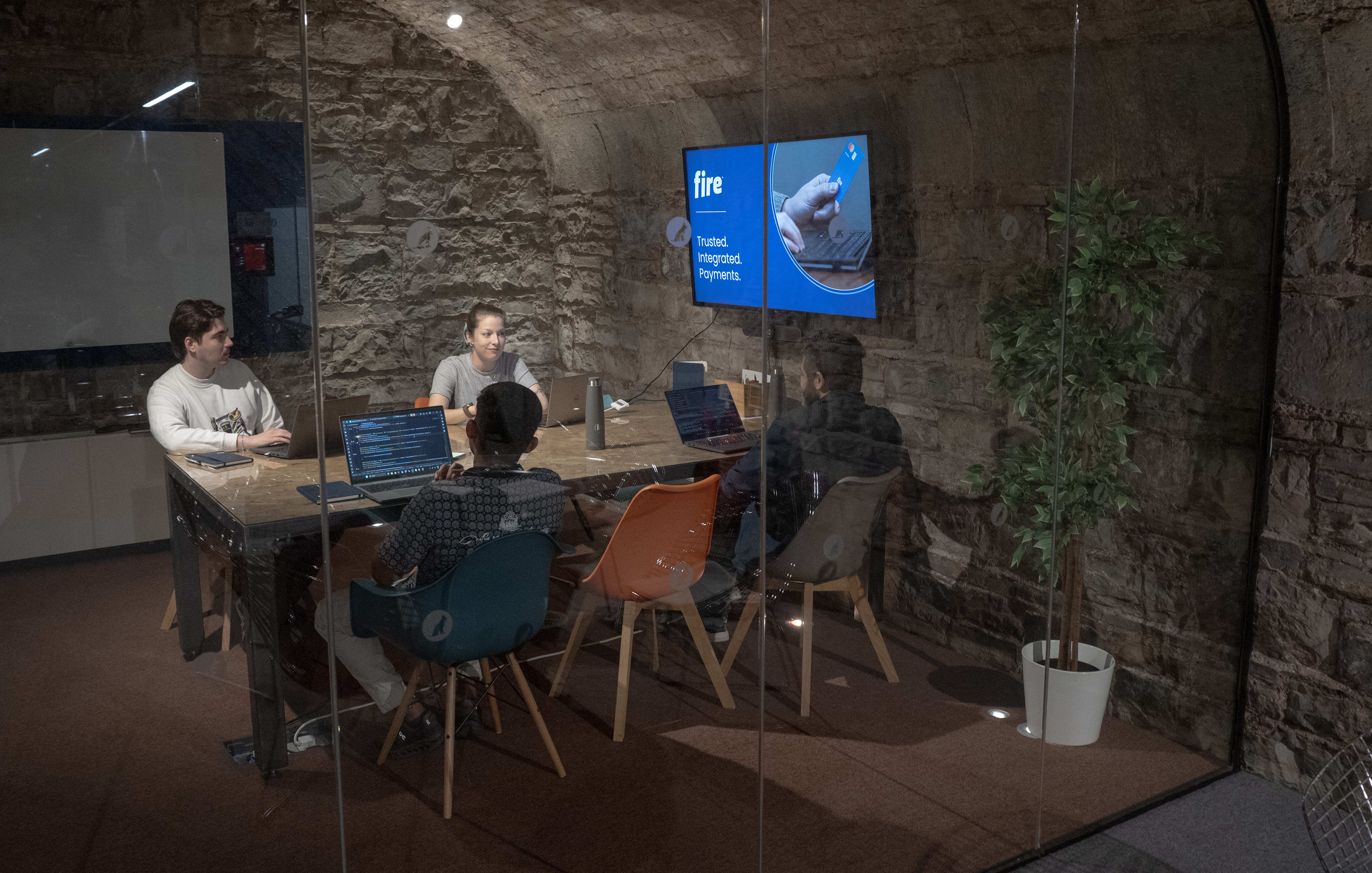 Four people sit around a table in a glass-walled meeting room with laptops, discussing in front of a screen displaying the Fire logo and the words “Trusted. Integrated. Payments.”