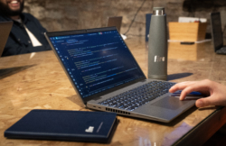 Close-up of a person working on a laptop showing lines of code on the screen, with a Fire-branded notebook and water bottle on the wooden table.