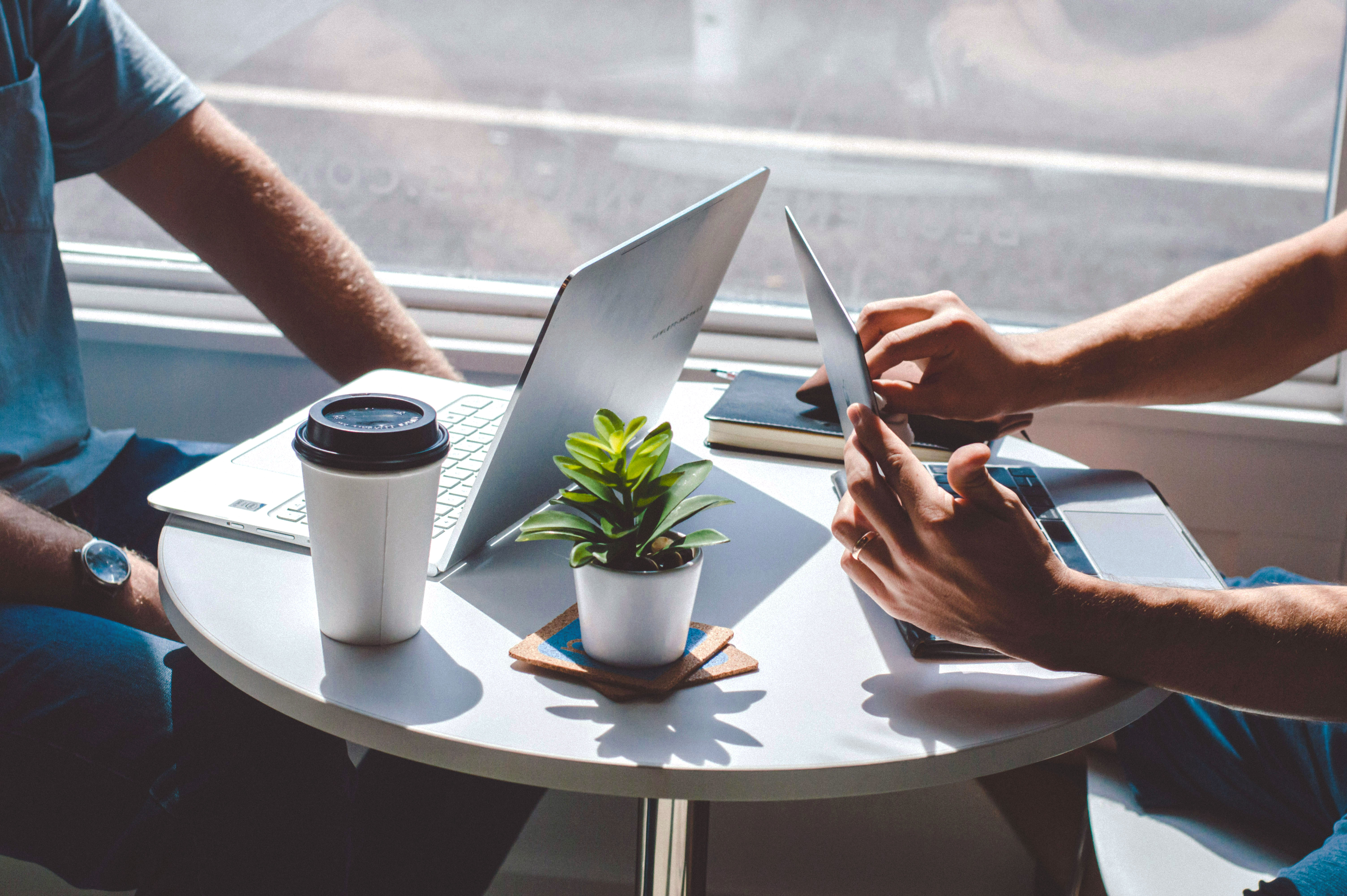 Two people sit at a small round table with laptops and a tablet, working together near a bright window. A takeaway coffee cup and a small potted plant are on the table.