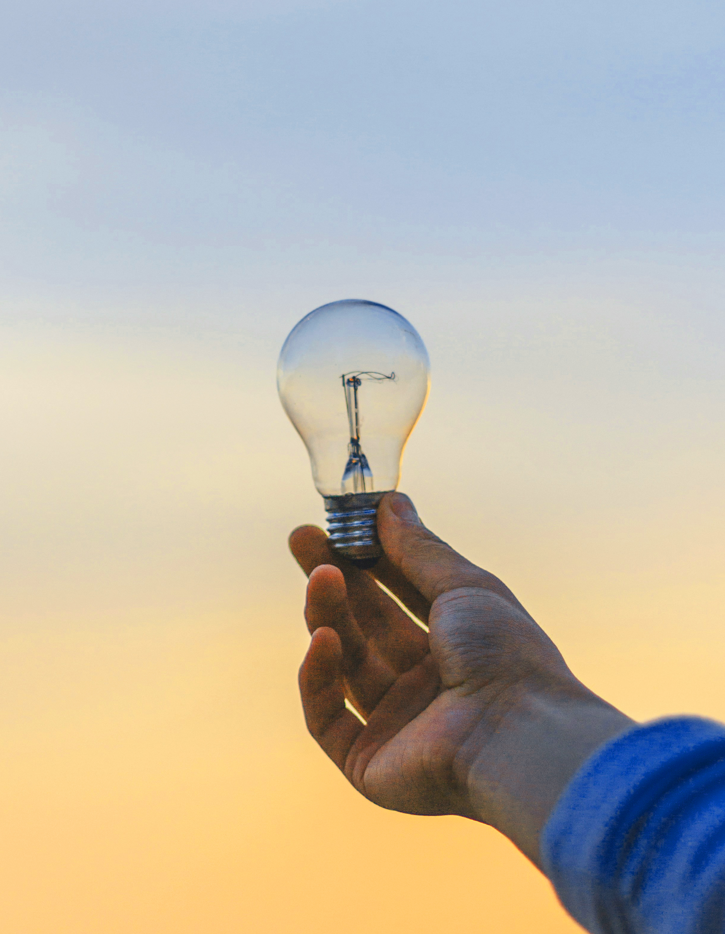 Hand holding a clear light bulb against a blue and golden sky.