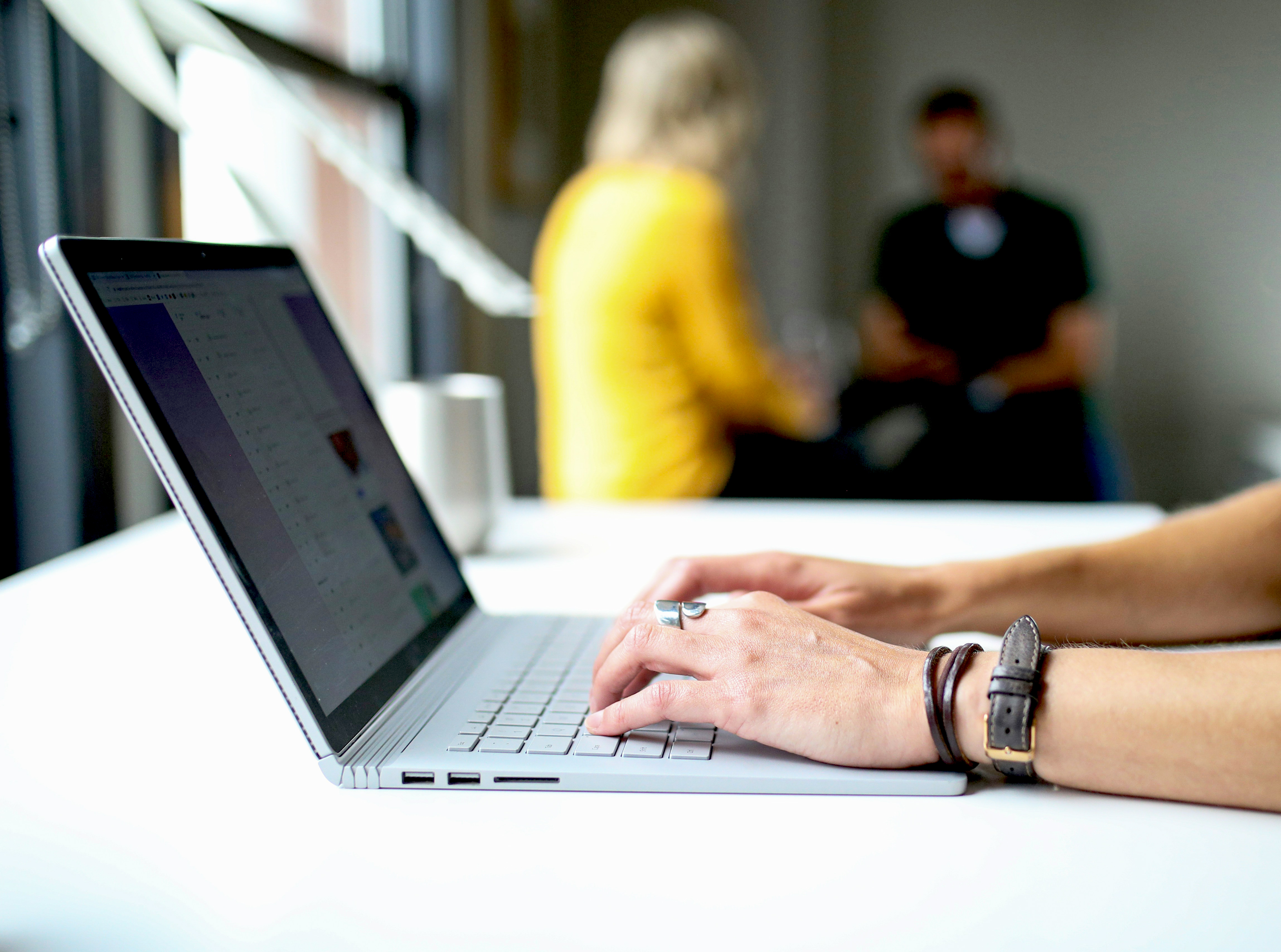 A person typing on a laptop at a table, with two people in the background.